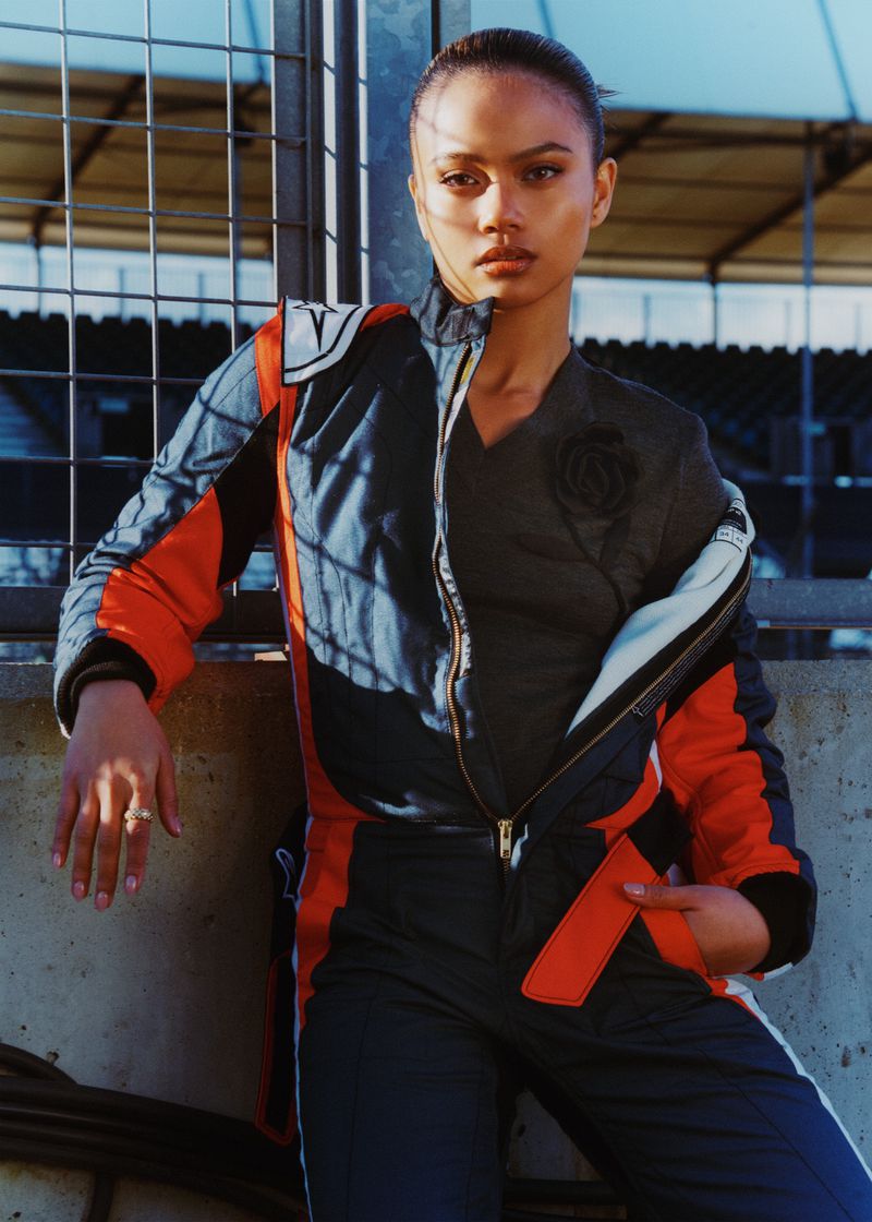 Woman with dark hair poses in racing jumpsuit against a metal fence and stadium seating backdrop under bright, direct sunlight.