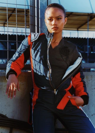 Woman with dark hair poses in racing jumpsuit against a metal fence and stadium seating backdrop under bright, direct sunlight.
