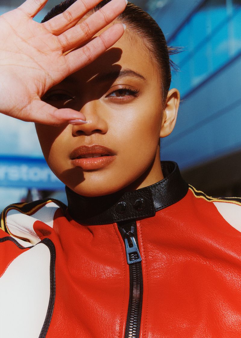 Portrait of a woman in a red leather jacket shielding her eyes with her hand against a blue window background, outdoor setting.