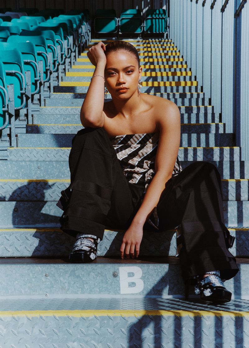 A young woman sits on stadium steps wearing a strapless top and dark pants, posing with her hand on her head in the daylight.