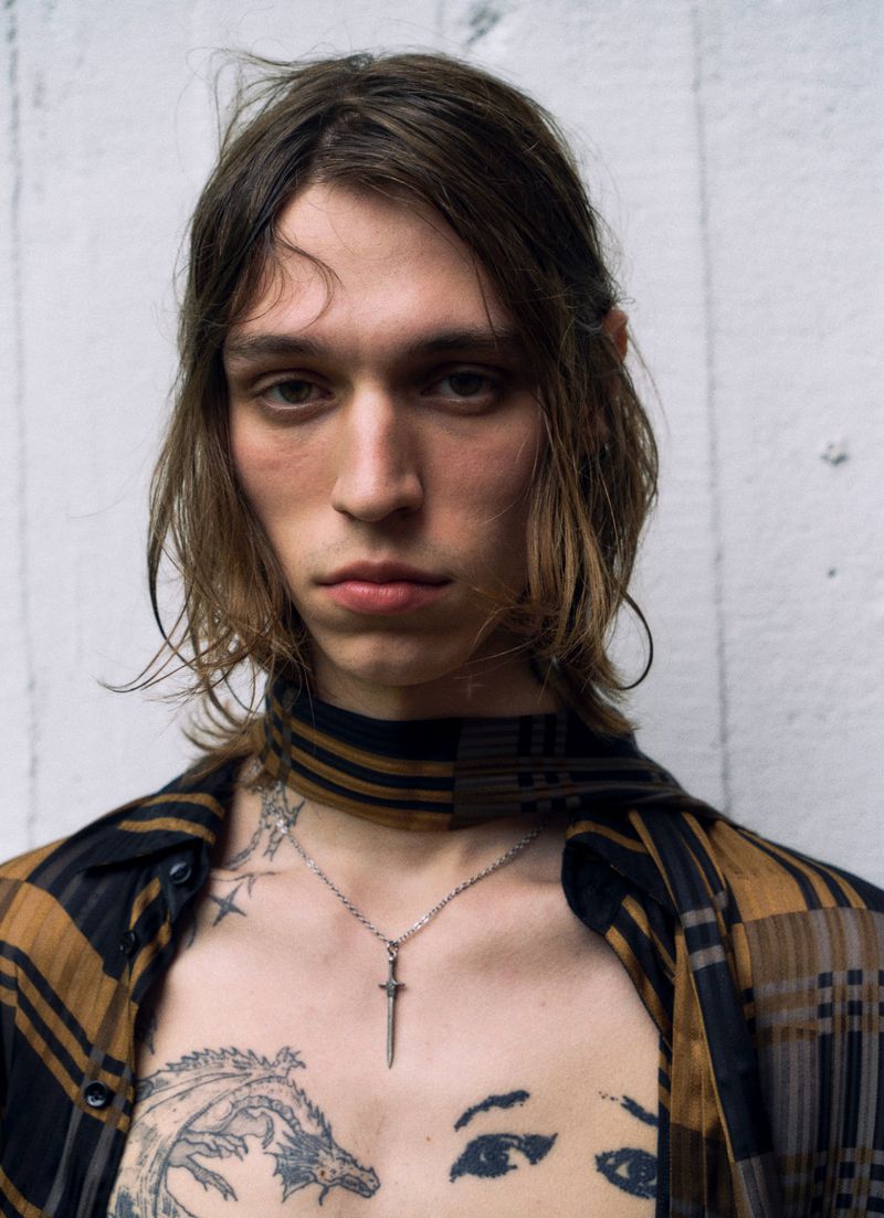 Portrait of a young adult male with brown hair, wearing a plaid shirt and necklace, with tattoos on his chest and face. 