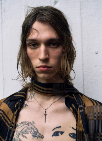 Portrait of a young adult male with brown hair, wearing a plaid shirt and necklace, with tattoos on his chest and face. 