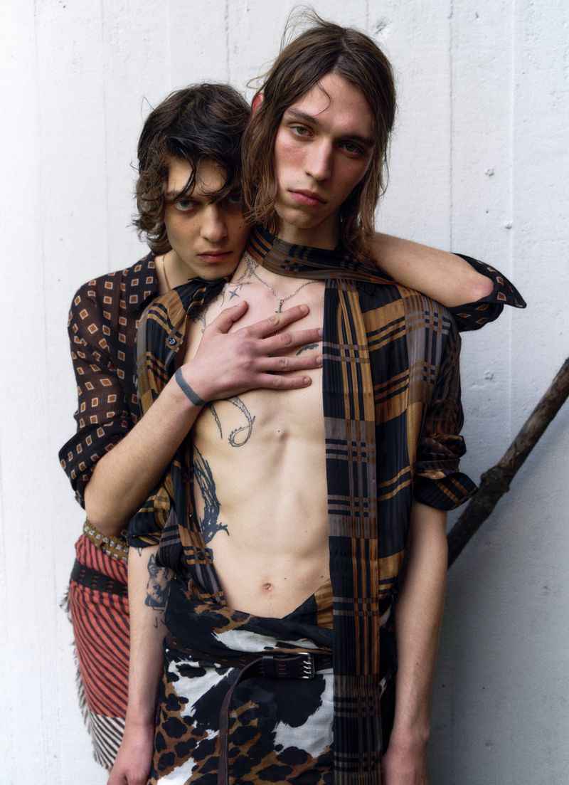Two adult men pose in casual wear against a neutral wall, one with hand on other's chest, showcasing tattoos and a unique fashion style.