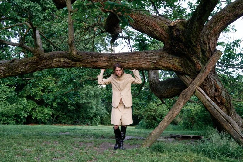 A person in a tan jacket and shorts stands beneath a large tree branch, wearing boots, in a grassy, forested park landscape.