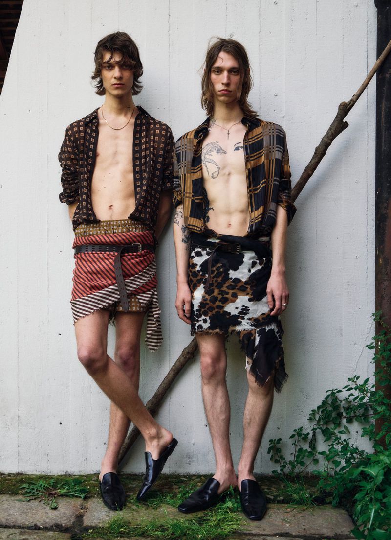 Two men pose outdoors with open shirts and wraps around their waist, showcasing fashion and style in a natural outdoor setting.
