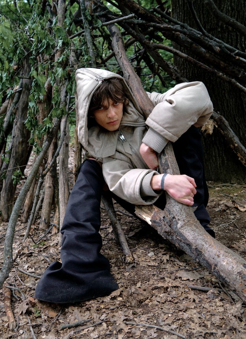 Man in hooded jacket sits among tree branches in a wooded environment. The scene features natural light and muted earth tone colors.