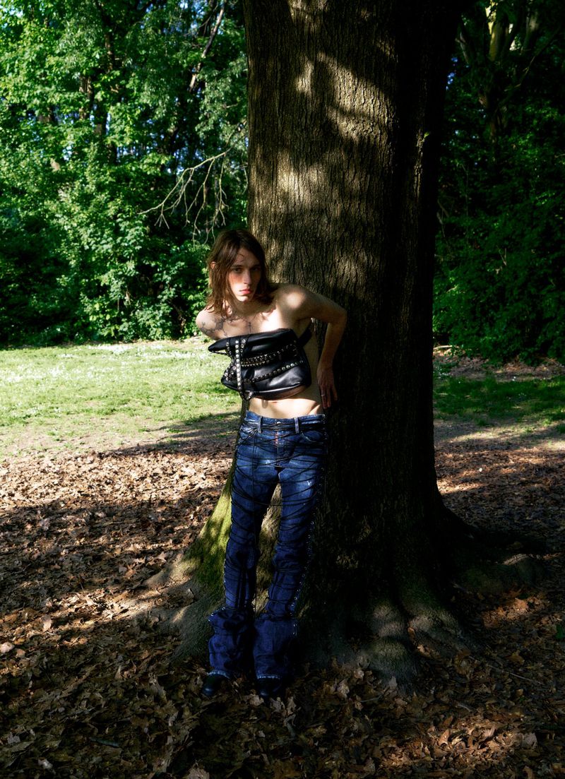 Adult female model poses near a tree in a leather bustier and jeans in an outdoor setting. Lush green foliage in the background.