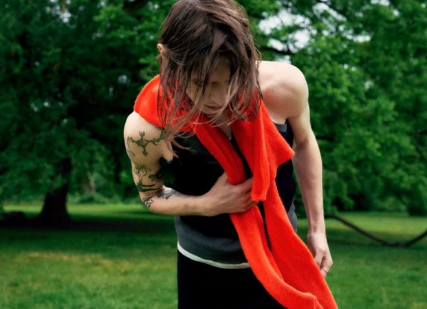 Adult male with tattoos outdoors in black tank top holding a red scarf in a green meadow surrounded by trees, captured in soft light.