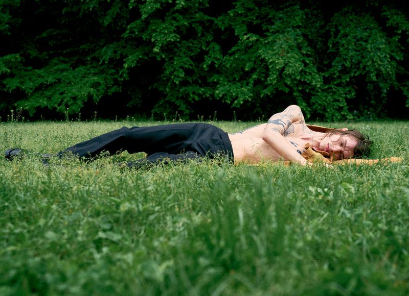 Tattooed person lying on a yellow blanket in a green grassy field with trees in the background, shirtless with dark pants.