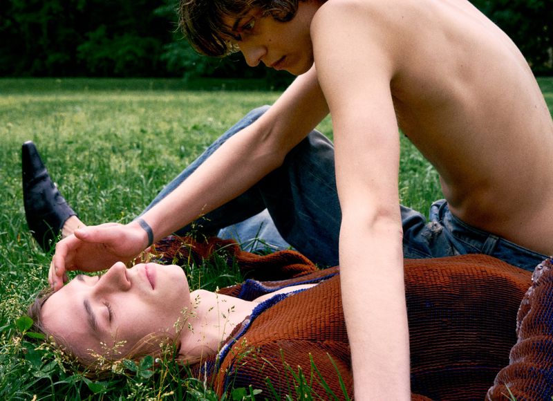 Two young adult men are lying on a grassy field, one with eyes closed and the other with a hand gently placed on the other's forehead.