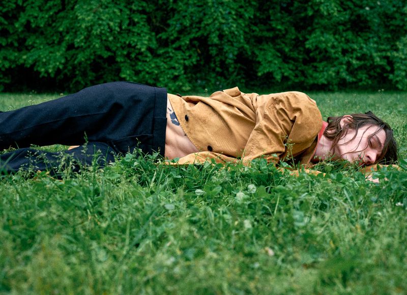 Young adult male with long hair resting on grass in an outdoor setting, wearing an open brown jacket and black pants, with green trees in the background.