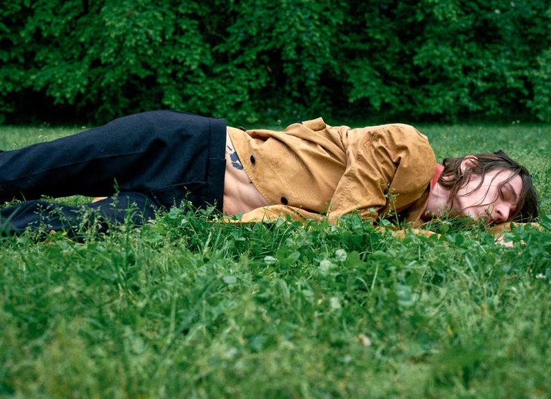 Young adult male with long hair resting on grass in an outdoor setting, wearing an open brown jacket and black pants, with green trees in the background.