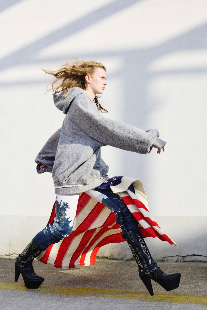 Fashionable woman strides confidently in American flag skirt, gray hoodie, and black heeled boots against a stark white wall.