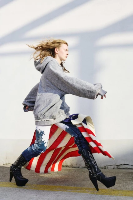 Fashionable woman strides confidently in American flag skirt, gray hoodie, and black heeled boots against a stark white wall.