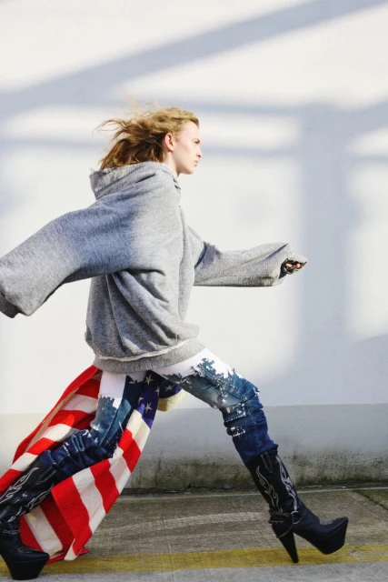 Fashion shot of a woman in a gray hoodie and patterned jeans with boots, carrying an American flag against a white wall.