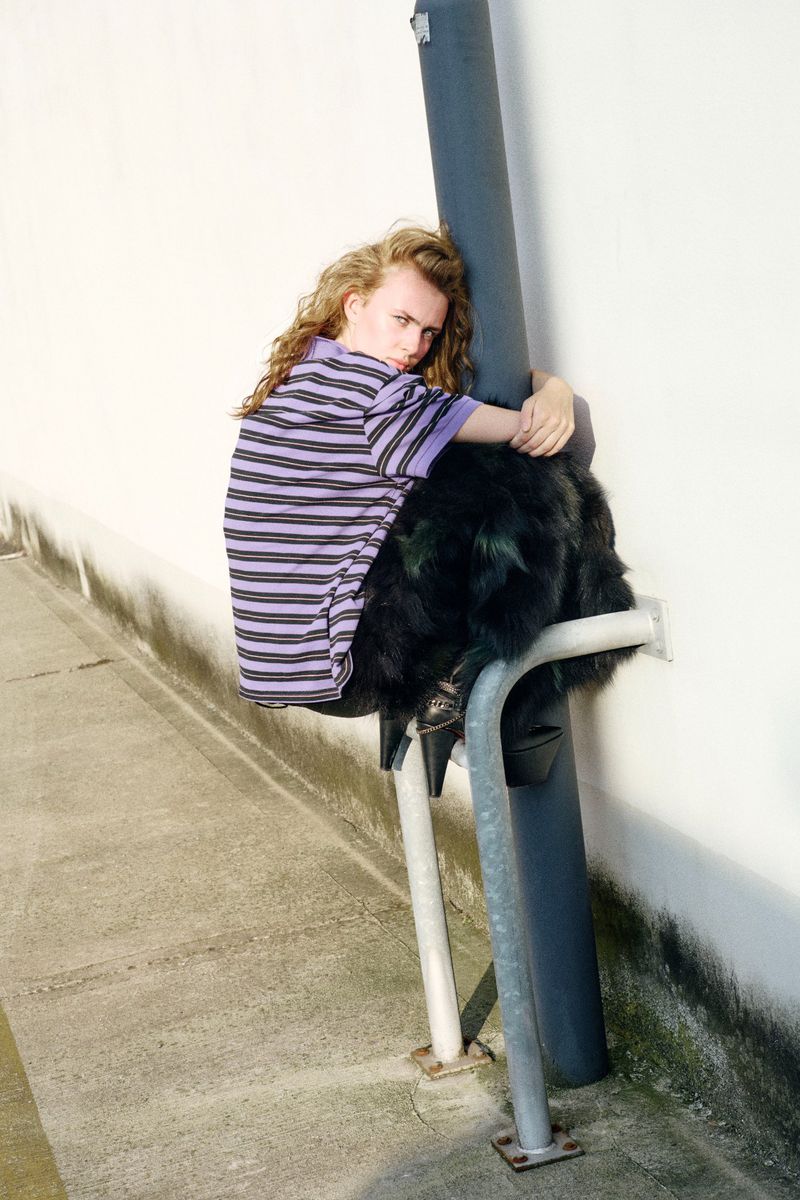 Adult with long wavy hair sits on metal structure outdoors. Person wears striped purple and black shirt with dark fur-like object.