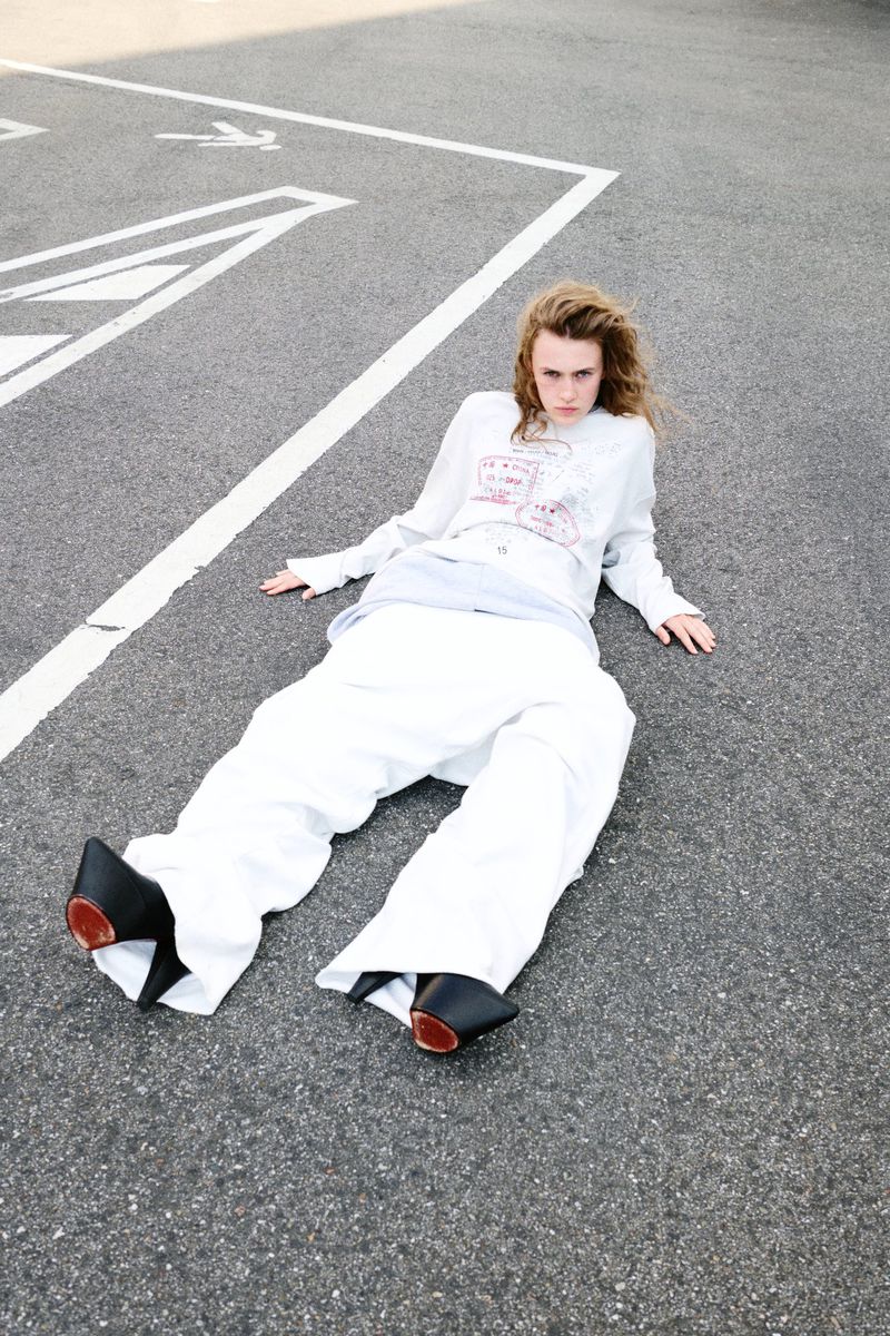 Young adult woman lying on asphalt wearing white pants and sweatshirt with graphic, black high heel shoes. Overhead, full shot.