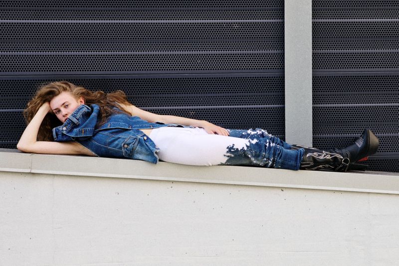 A young woman with long curly hair lies on a ledge in a blue denim jacket and pants with black western boots against a gray wall.