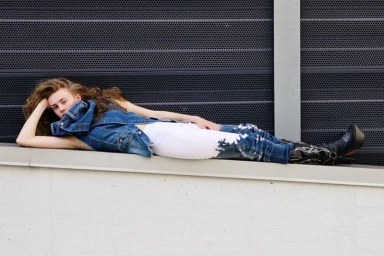 Woman lying horizontally on a concrete ledge wearing a denim vest, white and blue jeans, and black western boots.