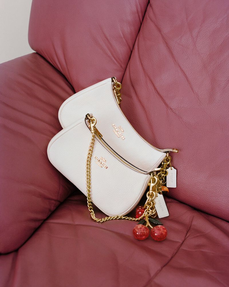 Two Coach bags in white leather with gold chains are displayed on a mauve leather surface, accented with cherry charms.