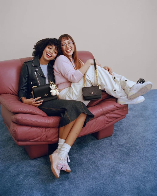 Two women smile on a couch, showcasing Coach purses. The woman on the left is in a leather jacket, while the other is in pink.