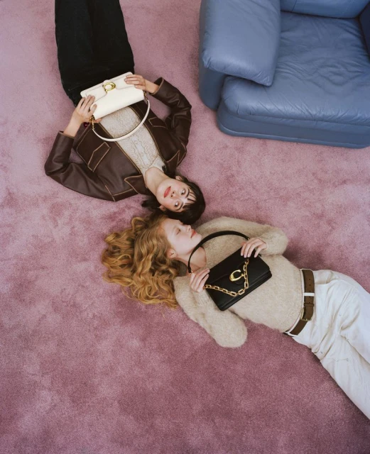 Two fashion models lie on a pink carpet with purses, wearing a brown jacket and beige sweater in a studio setting with a blue chair.