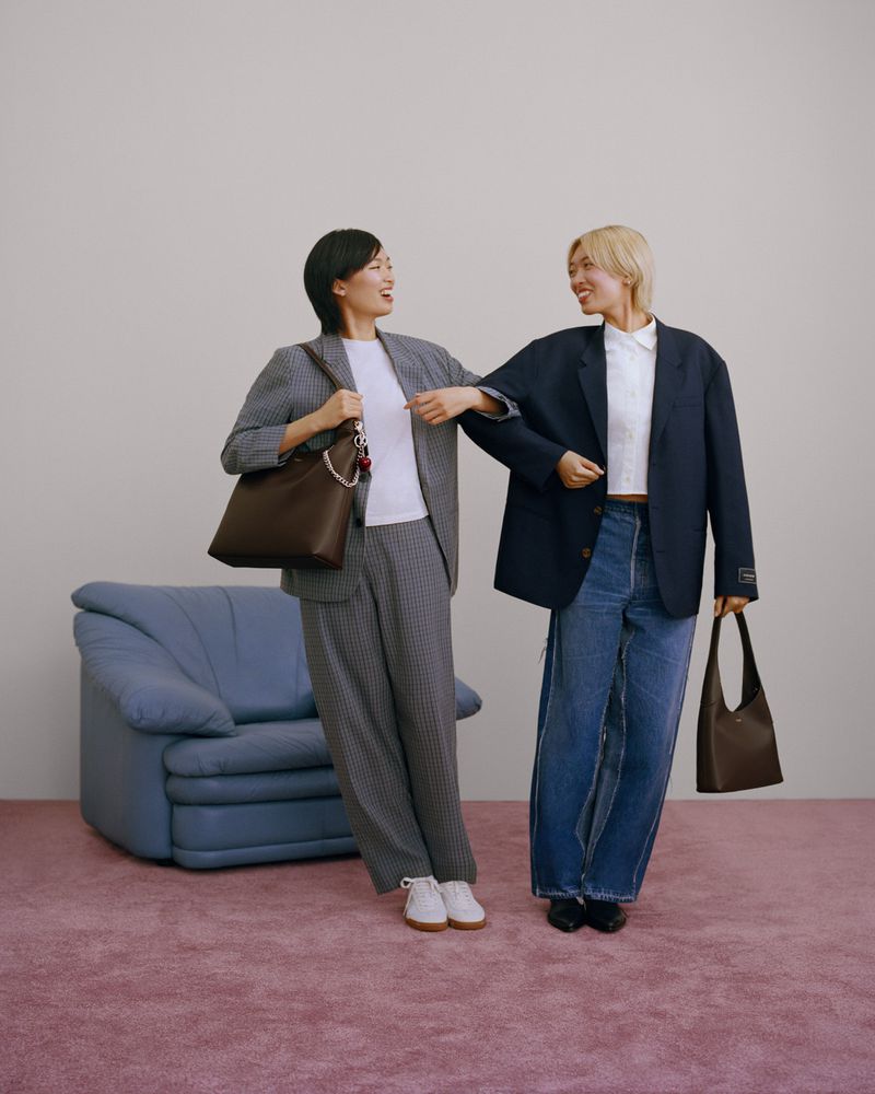 Two women in business-casual attire pose indoors with handbags. One wears a grey suit, the other jeans and a navy blazer.