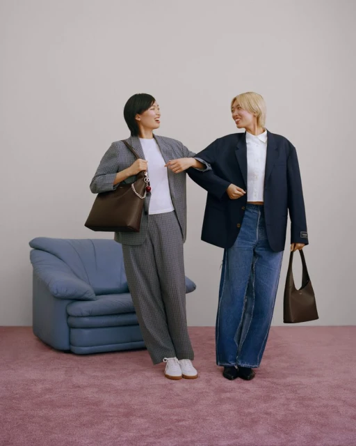 Two women in business-casual attire pose indoors with handbags. One wears a grey suit, the other jeans and a navy blazer.