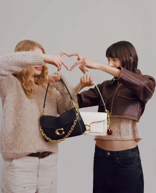 Two fashion models showcase Coach purses while forming a heart shape with their hands indoors, posing against a neutral backdrop.