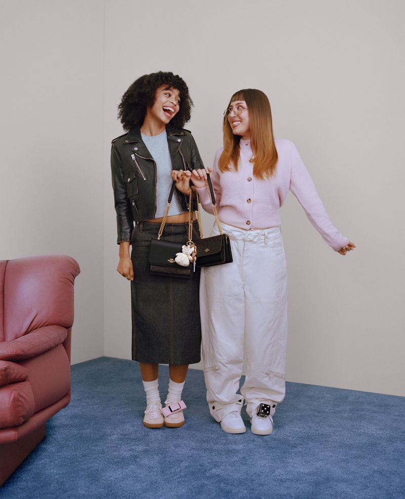 Two young women with purses pose indoors. One has curly hair, a black jacket, and a dark skirt. The other wears a pink cardigan.