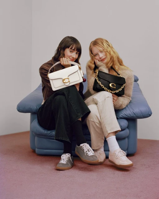 Two women pose with Coach purses in a studio setting. One holds a white purse, and the other holds a black purse with gold chain.