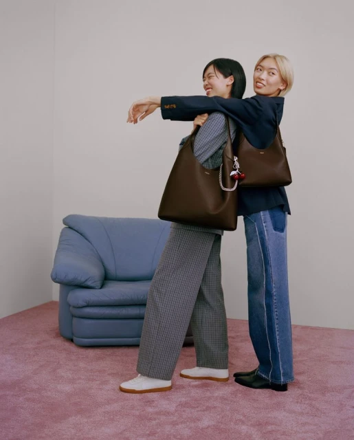 Two smiling Asian women wearing blazers and carrying leather tote bags pose in a pink-carpeted room with a blue couch.