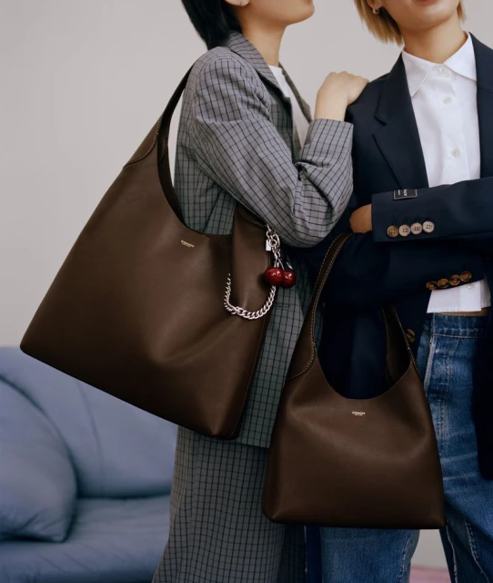 Two women pose indoors with matching brown leather shoulder bags and stylish blazers in a fashion editorial shot.