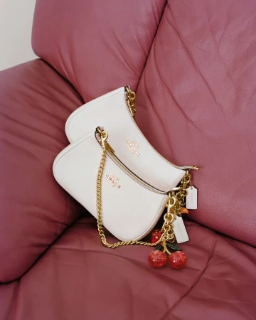 Two white leather Coach shoulder bags with gold hardware and a cherry charm resting on a mauve leather sofa.