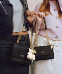 Two models standing side-by-side holding Coach chain shoulder bags and a white bear charm against a neutral studio backdrop.