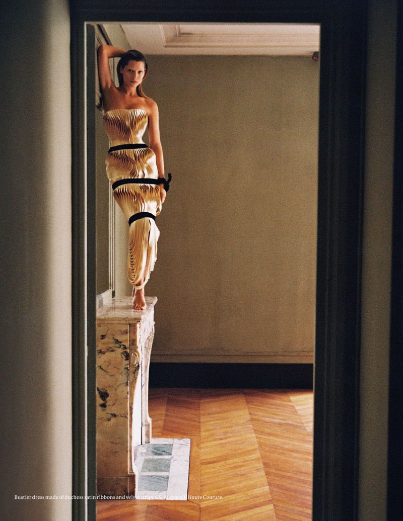Woman in high-fashion dress poses barefoot on a marble fireplace mantel, framed by a doorway, in a staged indoor setting with wood floor.