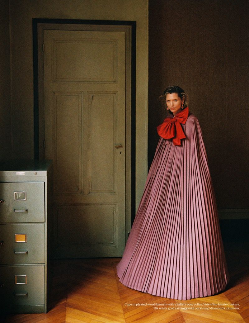 Woman in a pink pleated dress with a large red bow, standing on a parquet floor, next to a filing cabinet and a closed green door.