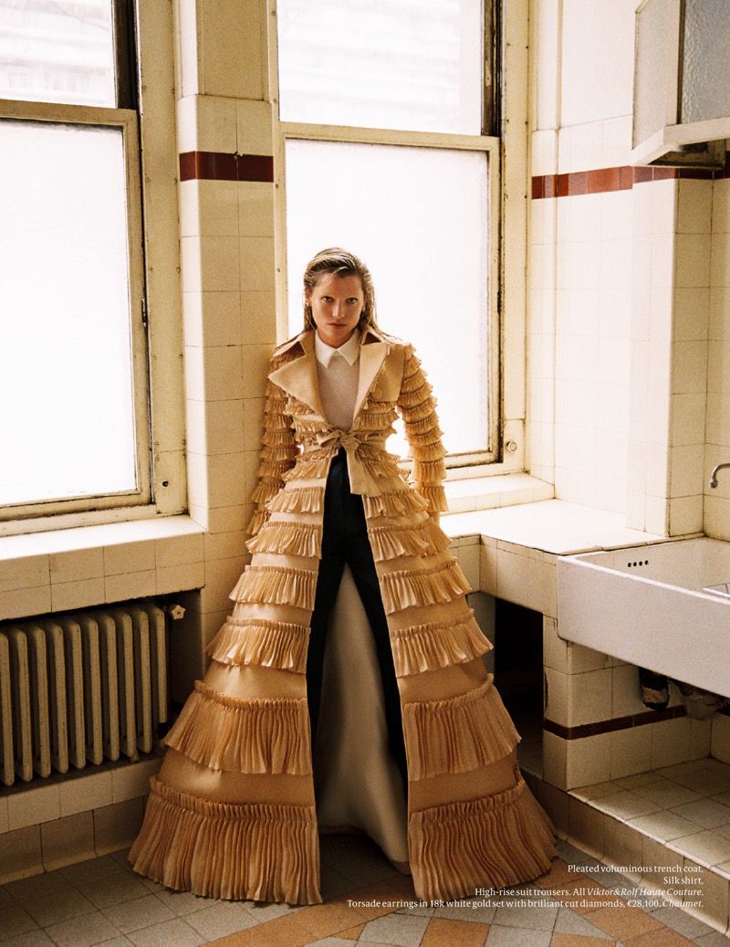 Fashion editorial shot of a female model in an ornate beige trench coat, black pants, and white blouse, posed in an indoor bathroom setting.