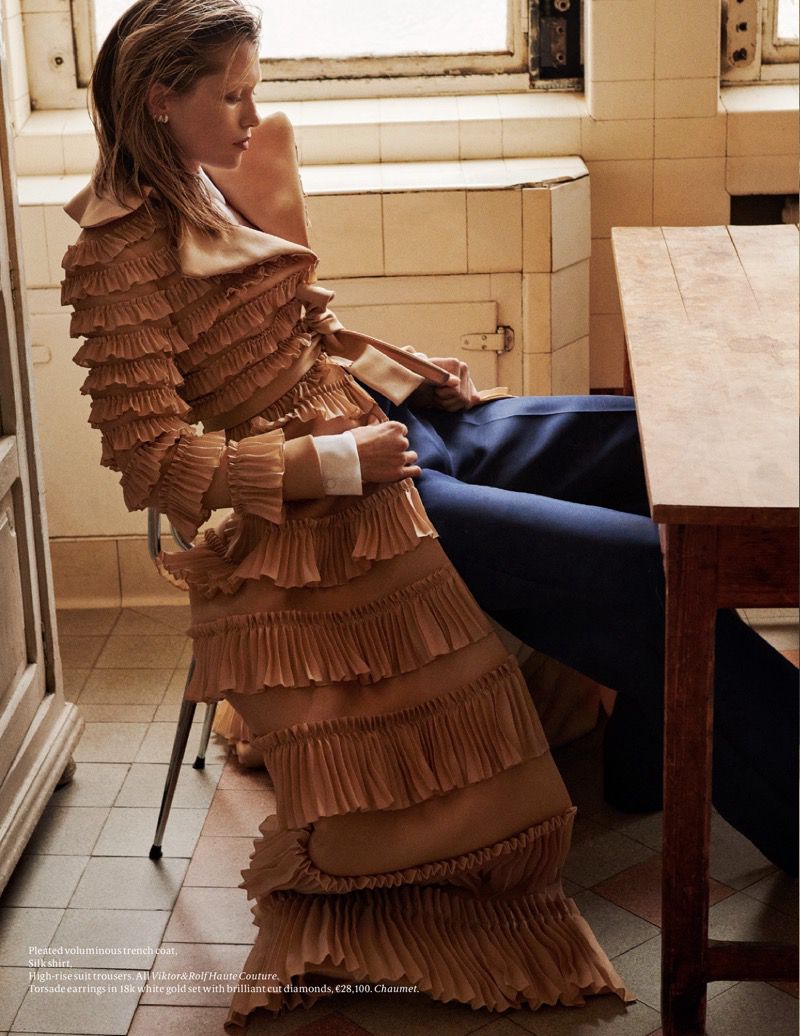 A woman in a peach-colored tiered dress is seated on a chair next to a wooden table with a tiled wall in the background.