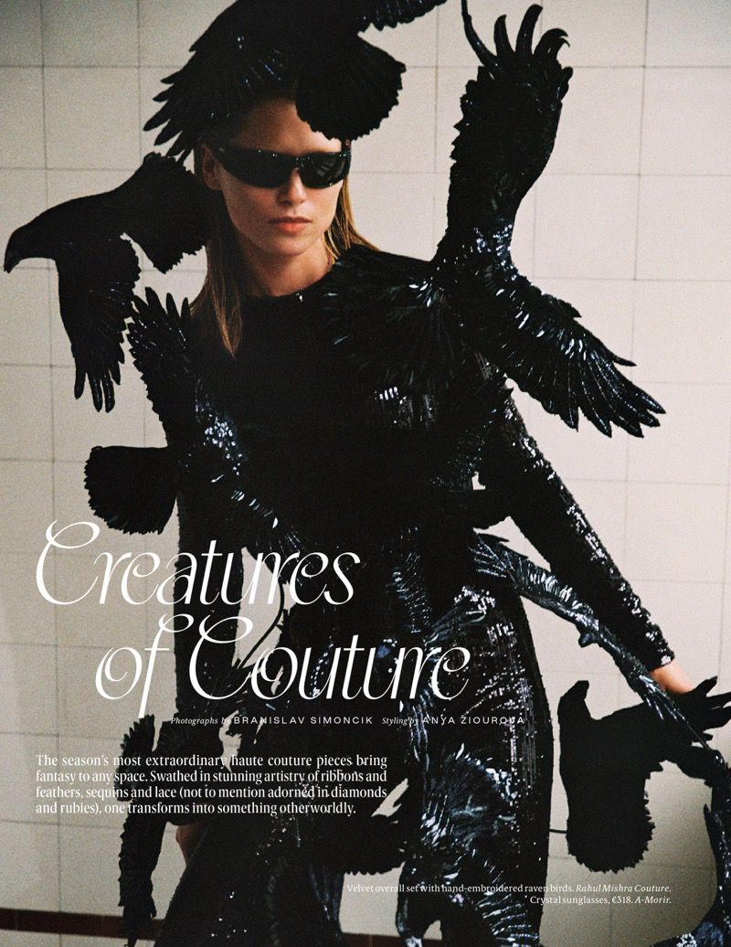 Editorial shot of a model in a black couture dress with bird-like adornments, wearing sunglasses, against a white tiled background.