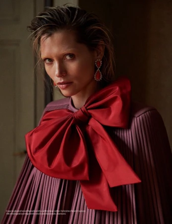 Portrait of an adult woman with fair skin wearing a lilac pleated dress with a large red bow and tear drop earrings indoors.