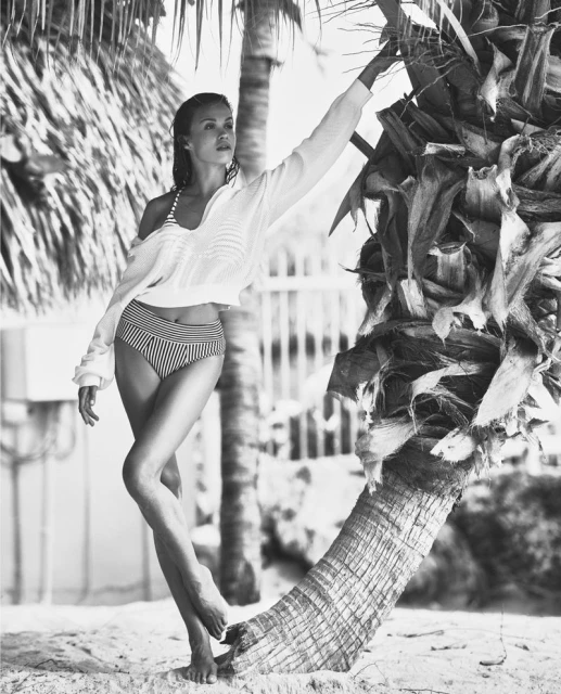Woman in striped bikini and white blouse poses near a palm tree on a sandy beach in a black and white lifestyle fashion shot.