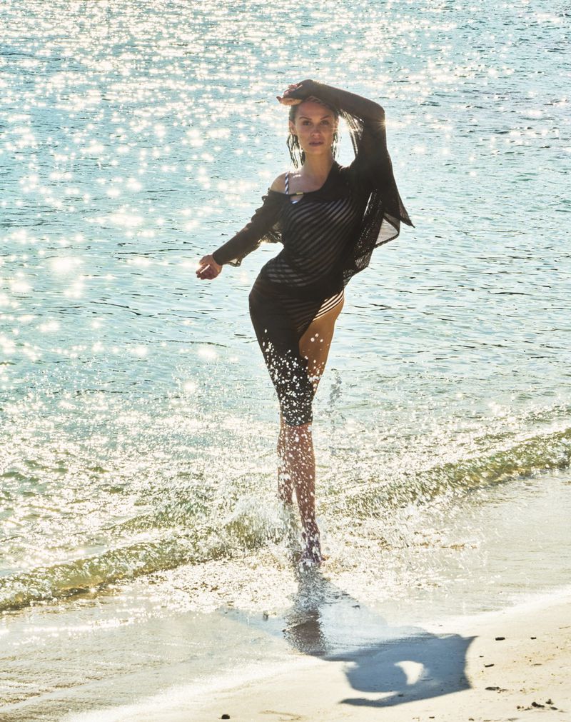 A woman in a black cover-up walks through the water on a sunny beach. The sunlight sparkles on the water creating a joyful summer vibe.