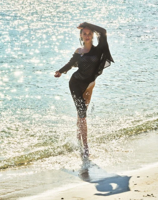 A woman in a black cover-up walks through the water on a sunny beach. The sunlight sparkles on the water creating a joyful summer vibe.
