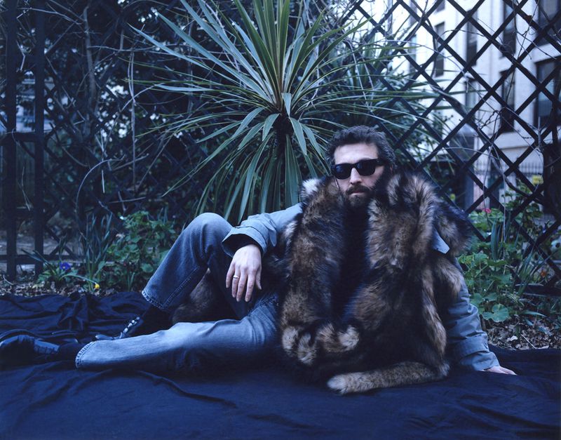 Man in sunglasses and fur coat reclines on dark fabric outdoors near tropical plant and wrought iron fence in daylight.