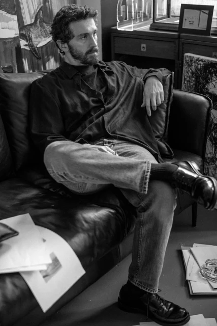 Black and white shot of an adult male with a beard and dark curly hair sitting on a black leather couch in a casual pose.