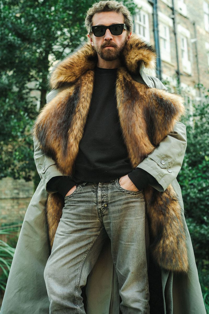 Man in sunglasses and fur-trimmed coat standing outdoors in front of tree and building, wearing jeans and a dark shirt.