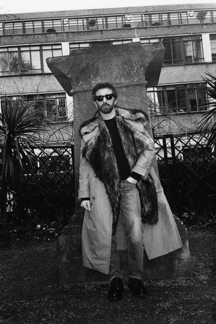 Man wearing fur-trimmed coat and sunglasses posing next to a concrete pillar in an urban garden with buildings in the background.