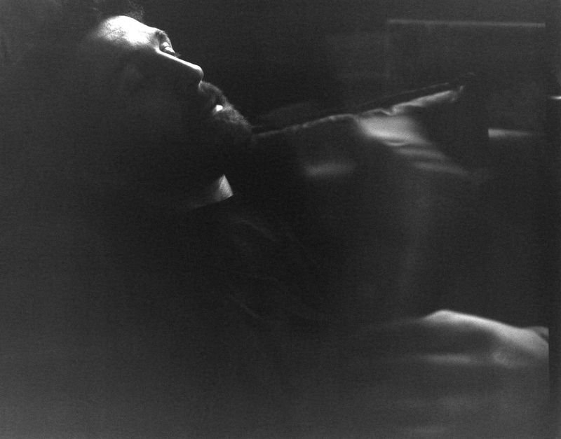 Black and white low-key portrait of a bearded man leaning back in deep shadow with light catching his facial profile.