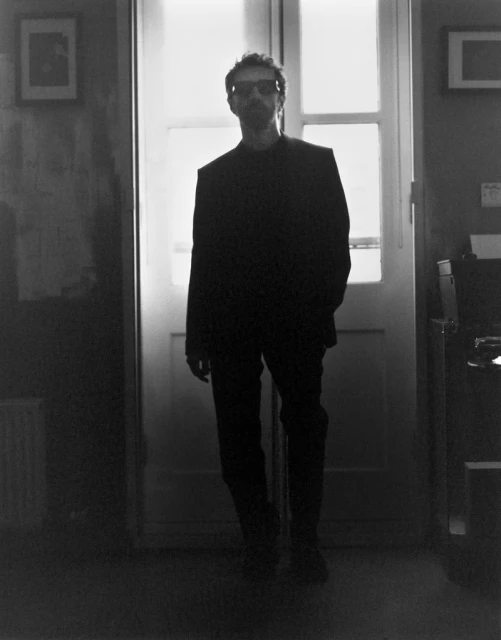 Man in a suit standing in a doorway, backlit by light. Interior with pictures on wall. Black and white aesthetic. Moody lighting.